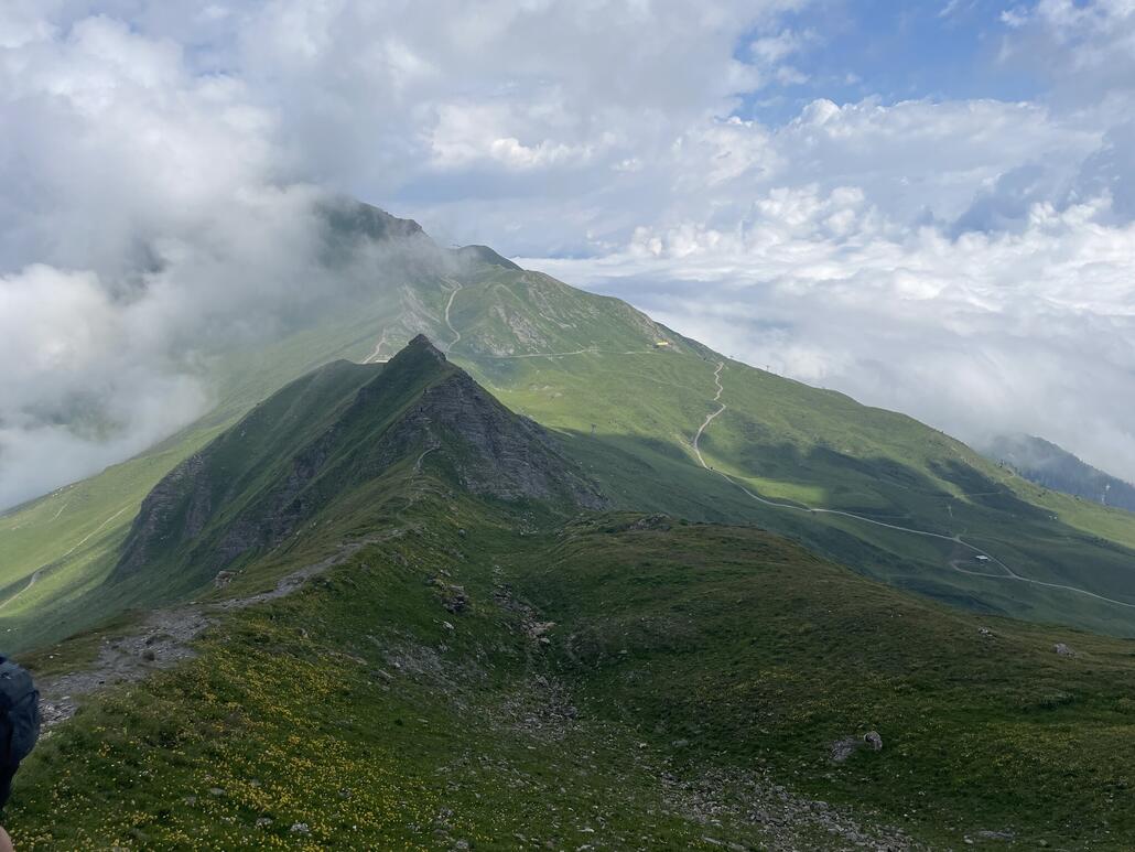 Wegverlauf vom Piz Danis Richtung Steinhaus. Das gelbe Gebäude in der Sone ist die Bergstation der Sesselbahn Cumacheals. Halb versteckt hinter dem einen Grat ist das Dach der Sesselbahnstation Lavoz zu erahnen.