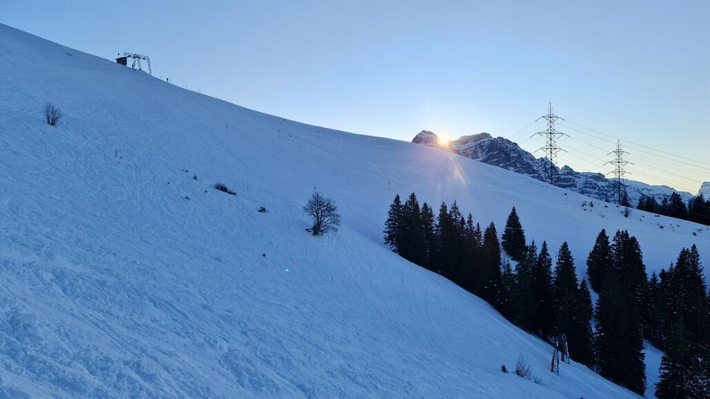 Die Sonne geht hinter den Glarner Gipfeln unter. Hier stehe ich auf der unpräparierten Abfahrtsroute, die im oberen Teil entlang des Schlepplifts verläuft und neben der schwarzen, präparierten Piste die einzige Variante von der Bergstation darstellt.